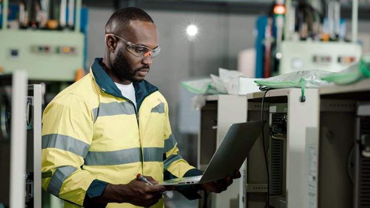 A man reads data on his laptop while standing between machines.