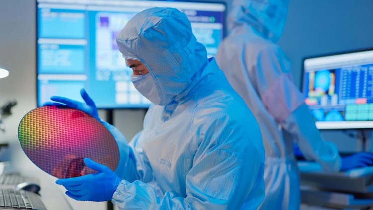 A technician in sterile coverall and gloves holds a wafer that reflects many different colors and checks it at a semiconductor manufacturing plant / Un technicien observe un wafer dans un centre de fabrication de semi-conducteurs