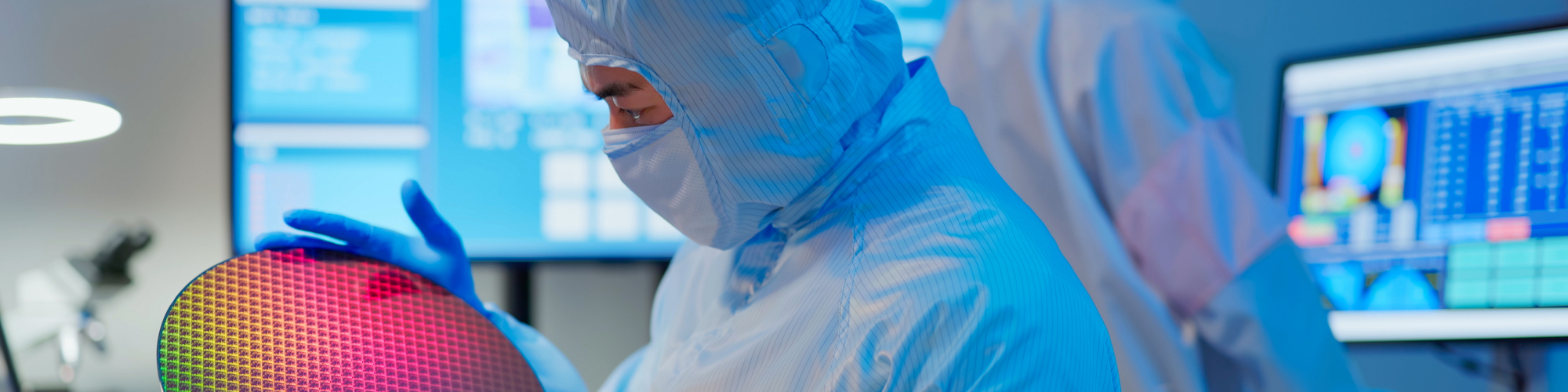 A technician in sterile coverall and gloves holds a wafer that reflects many different colors and checks it at a semiconductor manufacturing plant / Un technicien observe un wafer dans un centre de fabrication de semi-conducteurs
