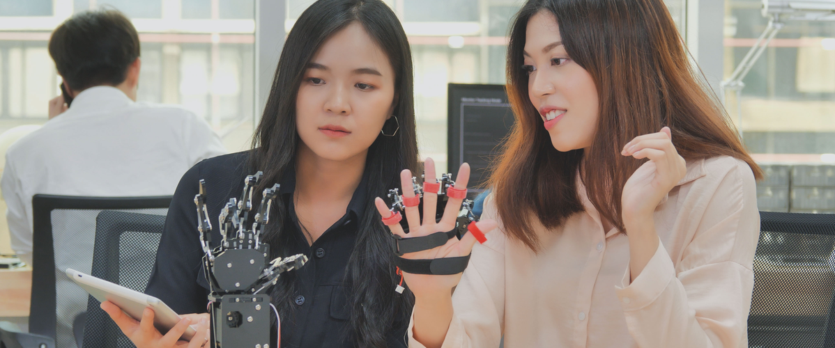 Two women sitting at a work table, one holding a tablet and the other displaying a robotic glove. The glove is equipped with sensors and mechanisms, showcasing a technological advancement in the field of robotics. In the background, a man is seated at a desk, talking on the phone.