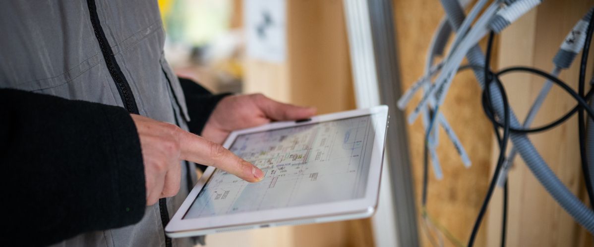 GettyImages - Main d'homme en gilet gris consulte une tablette sur un chantier, avec des câbles visibles en arrière-plan.