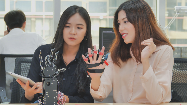 Two women sitting at a work table, one holding a tablet and the other displaying a robotic glove. The glove is equipped with sensors and mechanisms, showcasing a technological advancement in the field of robotics. In the background, a man is seated at a desk, talking on the phone.