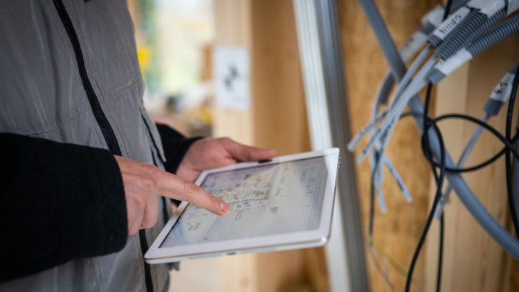 GettyImages - Main d'homme en gilet gris consulte une tablette sur un chantier, avec des câbles visibles en arrière-plan.