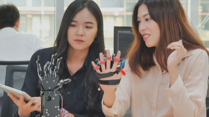 Two women sitting at a work table, one holding a tablet and the other displaying a robotic glove. The glove is equipped with sensors and mechanisms, showcasing a technological advancement in the field of robotics. In the background, a man is seated at a desk, talking on the phone.