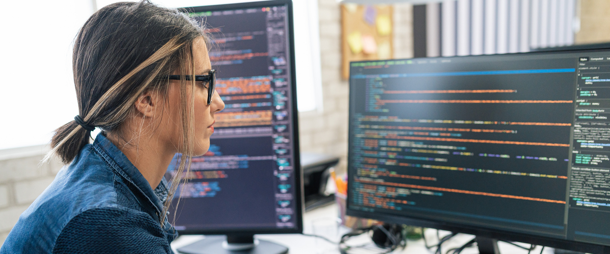 GettyImages - A woman is working on a computer, surrounded by two screens displaying code. The office environment is bright and modern, with stationery items visible on the desk.