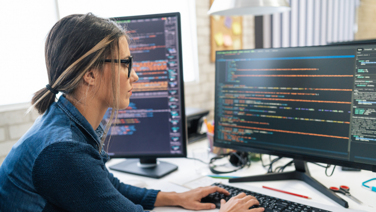 GettyImages - Une femme travaille sur un ordinateur, entourée de deux écrans affichant du code. L'environnement de bureau est lumineux et moderne, avec des éléments de papeterie visibles sur le bureau.