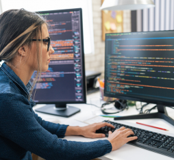 GettyImages - Une femme travaille sur un ordinateur, entourée de deux écrans affichant du code. L'environnement de bureau est lumineux et moderne, avec des éléments de papeterie visibles sur le bureau.