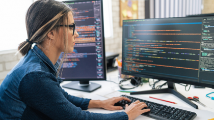 GettyImages - A woman is working on a computer, surrounded by two screens displaying code. The office environment is bright and modern, with stationery items visible on the desk.