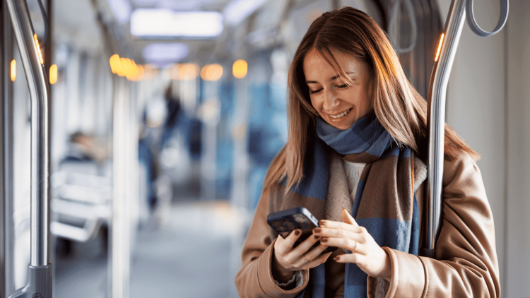 A woman stands in a train, holding a phone. She is wearing a beige coat and a blue and brown scarf. The interior of the train is bright, with seats and metal support bars.