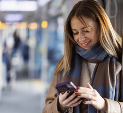 A woman stands in a train, holding a phone. She is wearing a beige coat and a blue and brown scarf. The interior of the train is bright, with seats and metal support bars.