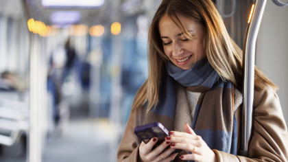 A woman stands in a train, holding a phone. She is wearing a beige coat and a blue and brown scarf. The interior of the train is bright, with seats and metal support bars.