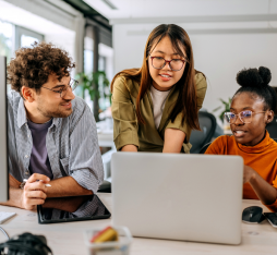 Trois personnes collaborent autour d'un ordinateur portable dans un environnement de bureau moderne. L'une d'elles, debout, explique quelque chose aux deux autres assis, qui semblent attentifs. Sur la table, on peut voir un ordinateur de bureau, une tablette et des accessoires de bureau. Des plantes et des bureaux sont visibles en arrière-plan.