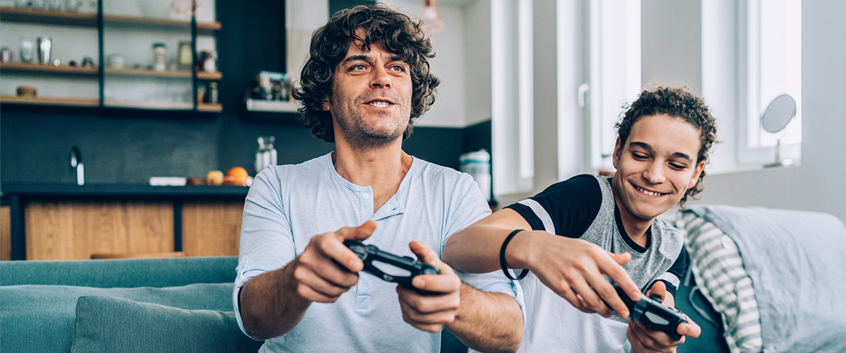 Two individuals sitting on a couch, playing video games. One is holding a game controller, focused on the screen, while the other appears to be giving advice or sharing strategies. The setting is modern and bright, with a kitchen visible in the background.