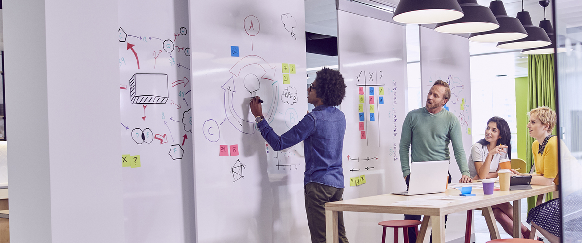 In a bright, modern office, a group of four people is engaged in a brainstorming session. A man is writing on a whiteboard, while three others, seated around a wooden table, are discussing and looking at a laptop. Colorful sticky notes are visible on the board. The space is decorated with colorful curtains and hanging lamps, creating a dynamic and collaborative atmosphere.