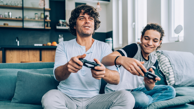 Two individuals sitting on a couch, playing video games. One is holding a game controller, focused on the screen, while the other appears to be giving advice or sharing strategies. The setting is modern and bright, with a kitchen visible in the background.