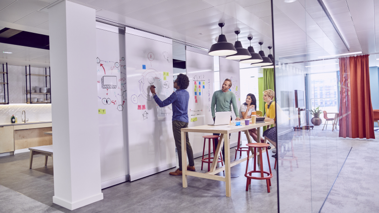 In a bright, modern office, a group of four people is engaged in a brainstorming session. A man is writing on a whiteboard, while three others, seated around a wooden table, are discussing and looking at a laptop. Colorful sticky notes are visible on the board. The space is decorated with colorful curtains and hanging lamps, creating a dynamic and collaborative atmosphere.