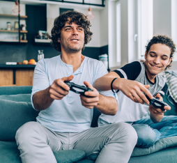 Deux personnes assises sur un canapé, jouant à des jeux vidéo. L'un tient une manette de jeu, concentré sur l'écran, tandis que l'autre semble donner des conseils ou partager des stratégies. L'environnement est moderne et lumineux, avec une cuisine visible en arrière-plan.