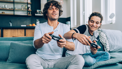 Two individuals sitting on a couch, playing video games. One is holding a game controller, focused on the screen, while the other appears to be giving advice or sharing strategies. The setting is modern and bright, with a kitchen visible in the background.