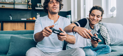 Deux personnes assises sur un canapé, jouant à des jeux vidéo. L'un tient une manette de jeu, concentré sur l'écran, tandis que l'autre semble donner des conseils ou partager des stratégies. L'environnement est moderne et lumineux, avec une cuisine visible en arrière-plan.