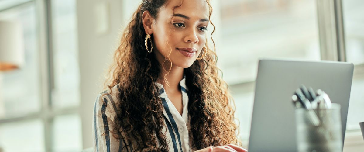 Une femme assise à un bureau, concentrée sur son ordinateur portable. Elle porte une chemise à rayures et a les cheveux longs et bouclés. Sur le bureau, on peut voir un carnet ouvert et un pot contenant des stylos. La lumière naturelle entre par une grande fenêtre, créant une ambiance chaleureuse et productive.