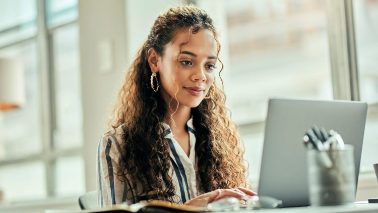 Une femme assise à un bureau, concentrée sur son ordinateur portable. Elle porte une chemise à rayures et a les cheveux longs et bouclés. Sur le bureau, on peut voir un carnet ouvert et un pot contenant des stylos. La lumière naturelle entre par une grande fenêtre, créant une ambiance chaleureuse et productive.