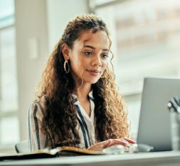 Une femme assise à un bureau, concentrée sur son ordinateur portable. Elle porte une chemise à rayures et a les cheveux longs et bouclés. Sur le bureau, on peut voir un carnet ouvert et un pot contenant des stylos. La lumière naturelle entre par une grande fenêtre, créant une ambiance chaleureuse et productive.