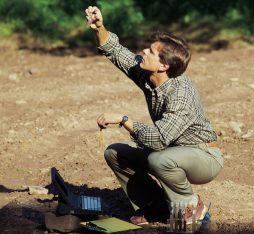 Un homme est accroupi sur un sol nu, tenant un objet en l'air avec une main et un crayon dans l'autre. À côté de lui, un ordinateur portable est ouvert, et il semble concentré sur son travail de recherche en extérieur.