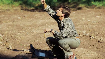 Un homme est accroupi sur un sol nu, tenant un objet en l'air avec une main et un crayon dans l'autre. À côté de lui, un ordinateur portable est ouvert, et il semble concentré sur son travail de recherche en extérieur.