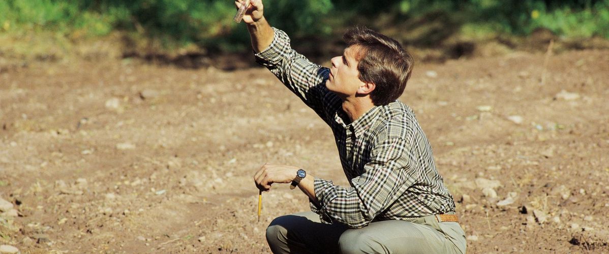 Un homme est accroupi sur un sol nu, tenant un objet en l'air avec une main et un crayon dans l'autre. À côté de lui, un ordinateur portable est ouvert, et il semble concentré sur son travail de recherche en extérieur.