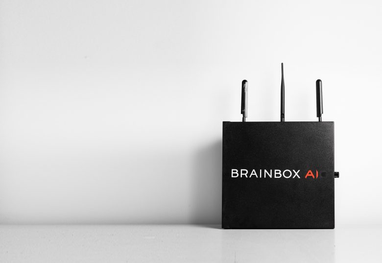 A black device named "BRAINBOX AI" is placed on a white surface. It has three antennas on top and is oriented to the right. The background is minimalist and plain.