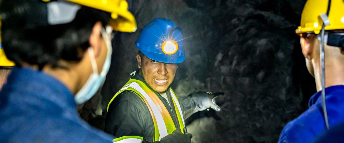 Un groupe de travailleurs en milieu minier écoute un collègue qui explique quelque chose. Ils portent des casques de sécurité jaunes et des masques. L'environnement est sombre, avec des murs rocheux visibles en arrière-plan. Le guide utilise une lampe frontale pour éclairer son chemin.