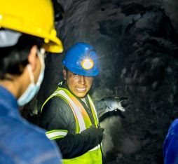 A group of mining workers is listening to a colleague who is explaining something. They are wearing yellow safety helmets and masks. The environment is dark, with rocky walls visible in the background. The guide is using a headlamp to light his way.