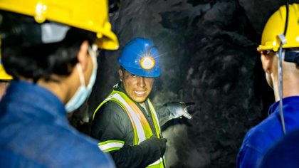 A group of mining workers is listening to a colleague who is explaining something. They are wearing yellow safety helmets and masks. The environment is dark, with rocky walls visible in the background. The guide is using a headlamp to light his way.