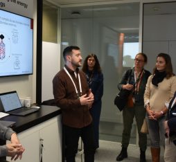 Deux présentateurs expliquent des concepts liés à BrainBox AI devant un écran affichant des graphiques et des informations. Six visiteurs se tiennent debout, attentifs.