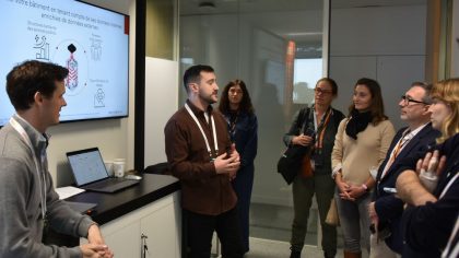 Deux présentateurs expliquent des concepts liés à BrainBox AI devant un écran affichant des graphiques et des informations. Six visiteurs se tiennent debout, attentifs.