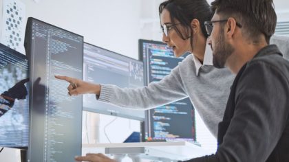 Two people collaborate in front of computer screens, one pointing something out to the other. The screens display computer code in a modern office environment.