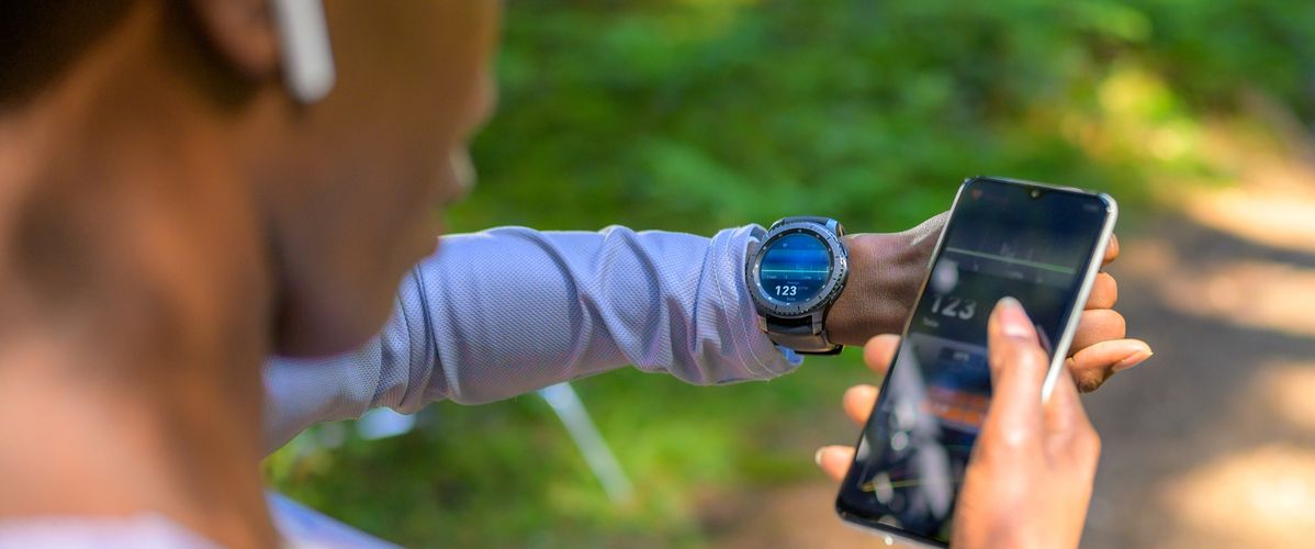 Une personne vérifie son rythme cardiaque sur une montre connectée tout en tenant un smartphone. L'environnement est naturel, avec des arbres en arrière-plan.