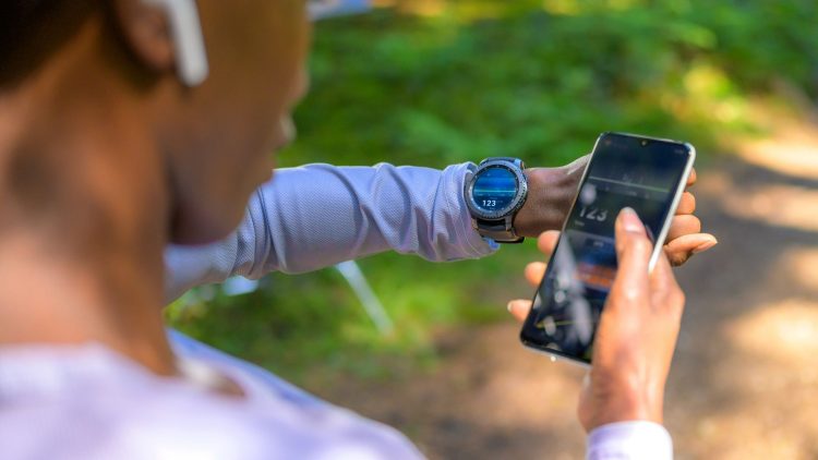 Une personne vérifie son rythme cardiaque sur une montre connectée tout en tenant un smartphone. L'environnement est naturel, avec des arbres en arrière-plan.