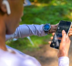 Une personne vérifie son rythme cardiaque sur une montre connectée tout en tenant un smartphone. L'environnement est naturel, avec des arbres en arrière-plan.