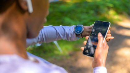 Une personne vérifie son rythme cardiaque sur une montre connectée tout en tenant un smartphone. L'environnement est naturel, avec des arbres en arrière-plan.