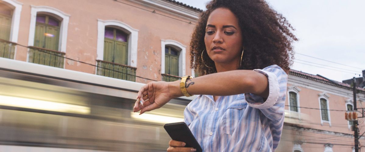 Une femme avec des cheveux bouclés se tient sur un trottoir, regardant sa montre tout en tenant un téléphone dans sa main. En arrière-plan, un tramway passe rapidement, flou, tandis que des bâtiments anciens avec des fenêtres vertes sont visibles.