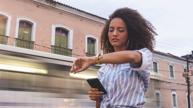 Une femme avec des cheveux bouclés se tient sur un trottoir, regardant sa montre tout en tenant un téléphone dans sa main. En arrière-plan, un tramway passe rapidement, flou, tandis que des bâtiments anciens avec des fenêtres vertes sont visibles.