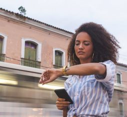 Une femme avec des cheveux bouclés se tient sur un trottoir, regardant sa montre tout en tenant un téléphone dans sa main. En arrière-plan, un tramway passe rapidement, flou, tandis que des bâtiments anciens avec des fenêtres vertes sont visibles.