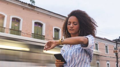 Une femme avec des cheveux bouclés se tient sur un trottoir, regardant sa montre tout en tenant un téléphone dans sa main. En arrière-plan, un tramway passe rapidement, flou, tandis que des bâtiments anciens avec des fenêtres vertes sont visibles.