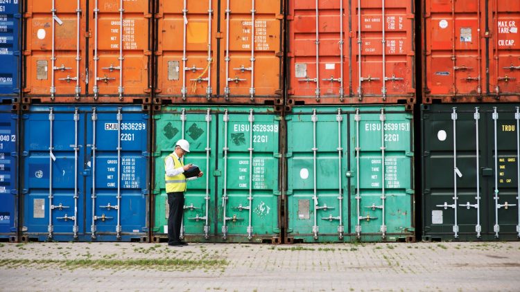 A man in a safety vest reviews documents in front of a row of colorful shipping containers at a port.