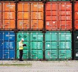 A man in a safety vest reviews documents in front of a row of colorful shipping containers at a port.