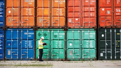 Un homme en gilet de sécurité examine des documents devant une rangée de conteneurs de transport colorés dans un port.