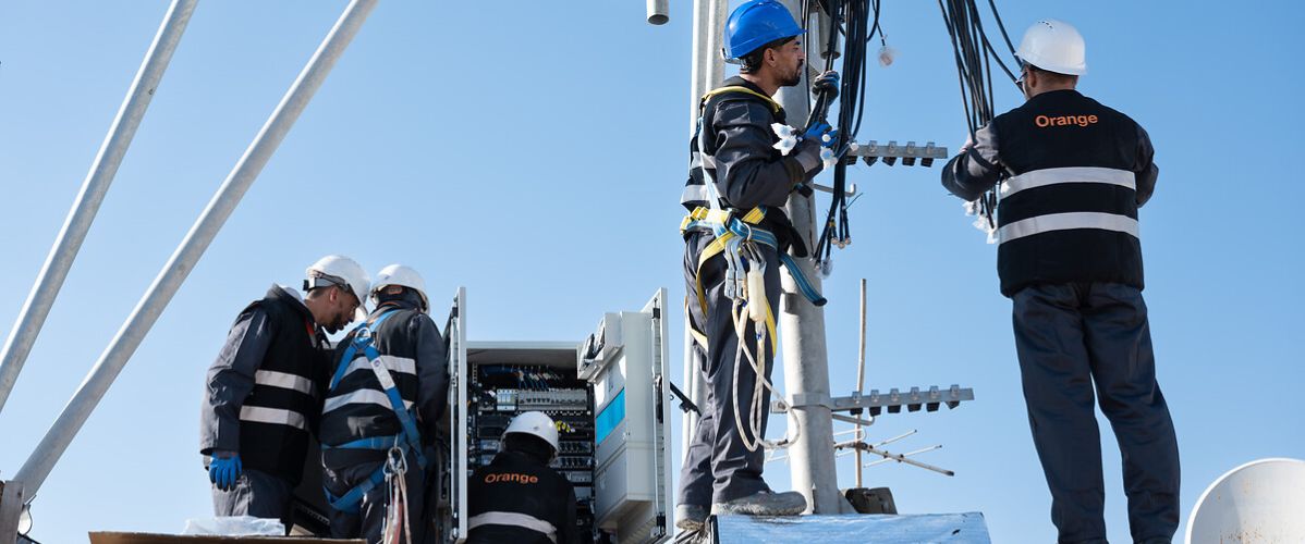 L'image montre un groupe de techniciens travaillant sur une tour de télécommunications. Ils sont engagés dans des tâches d'installation ou de maintenance, portant des casques de sécurité et des harnais, ce qui souligne l'importance de la sécurité.