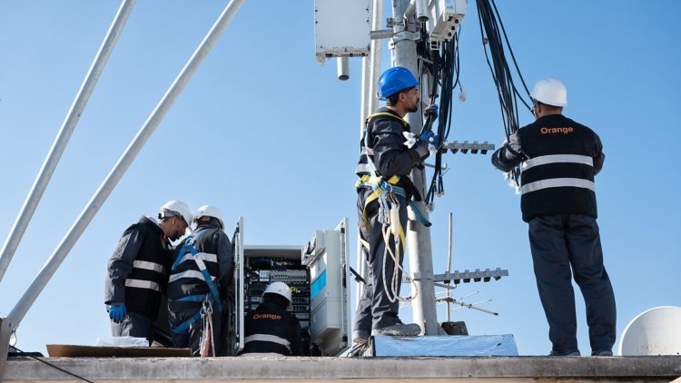 The image depicts a group of technicians working on a telecommunications tower. They are engaged in installation or maintenance tasks, wearing safety helmets and harnesses, highlighting a focus on safety. The presence of equipment suggests they are likely involved in enhancing or troubleshooting network services.