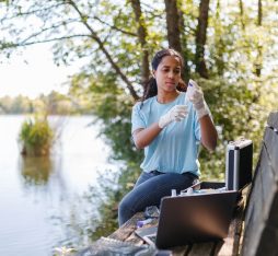 Young woman wearing gloves conducts environmental research by a lake. She uses equipment including a laptop and test kits. Trees and water in the background.